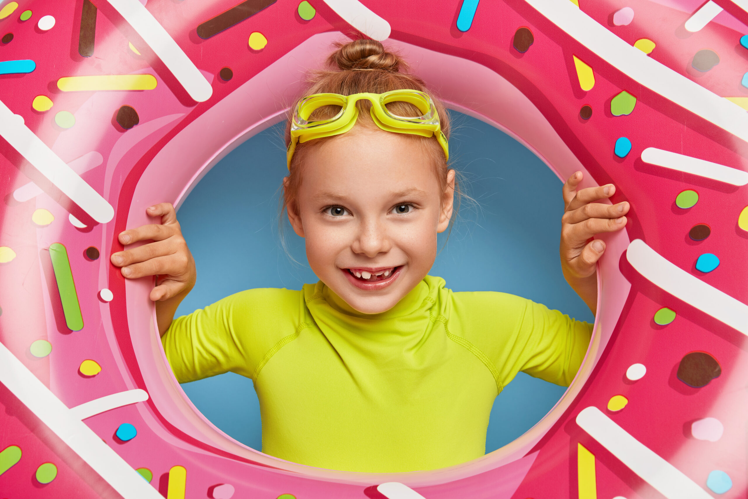 Close up shot of cute small kid wears goggles, swimwear, looks through rubber swimring, swims in sea, poses at beach alone, lives healthy life, smiles broadly. Children, summer, rest concept